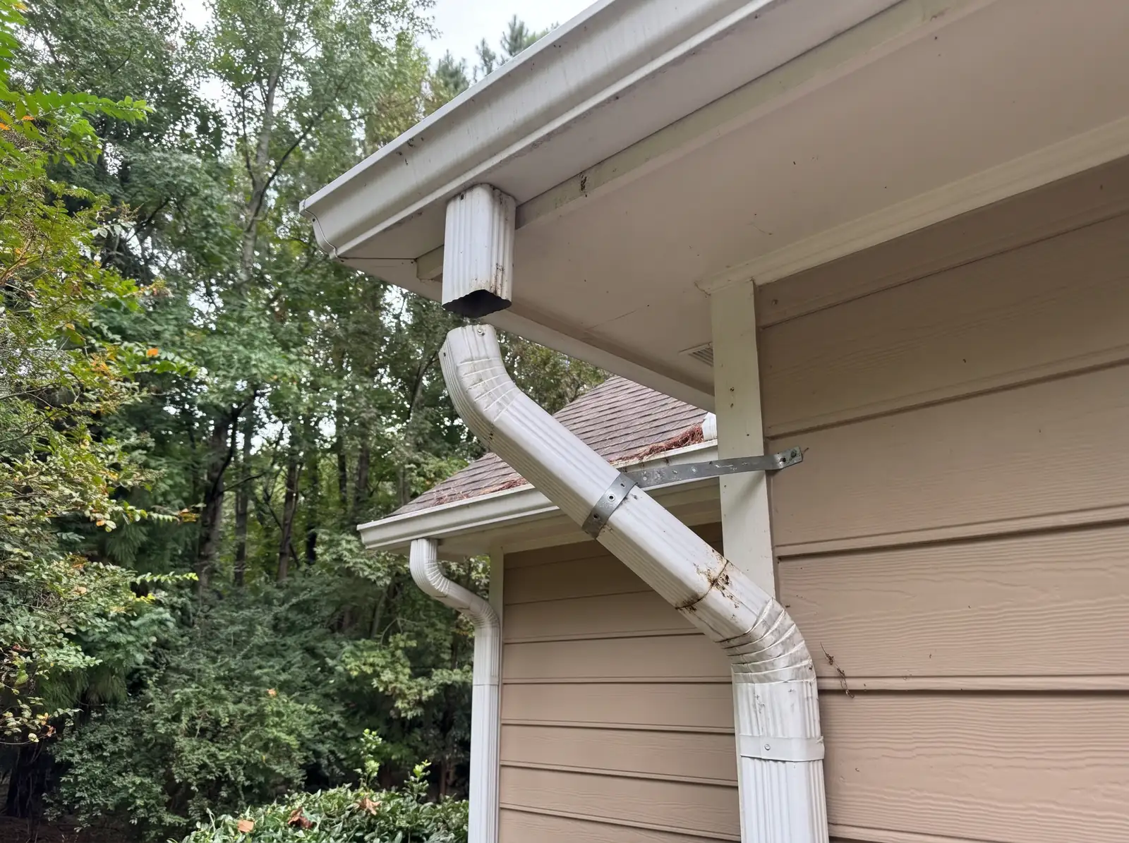 White downspout running at an angle from the eave, held to tan siding by a single strap
